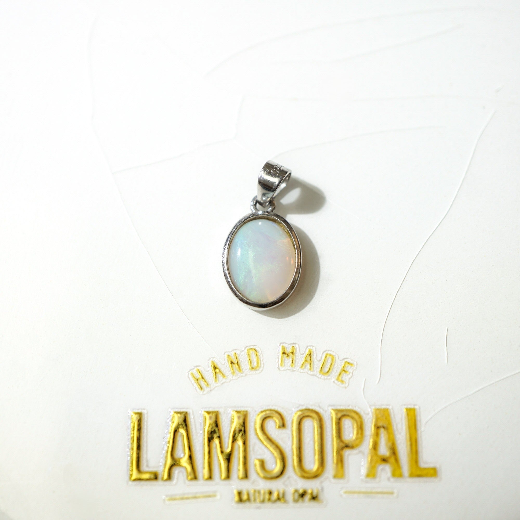 A silver oval pendant with a milky white iridescent opal stone, displayed on a white textured background with gold foil branding that reads 'HAND MADE LAMSOPAL NATURAL OPAL'.
