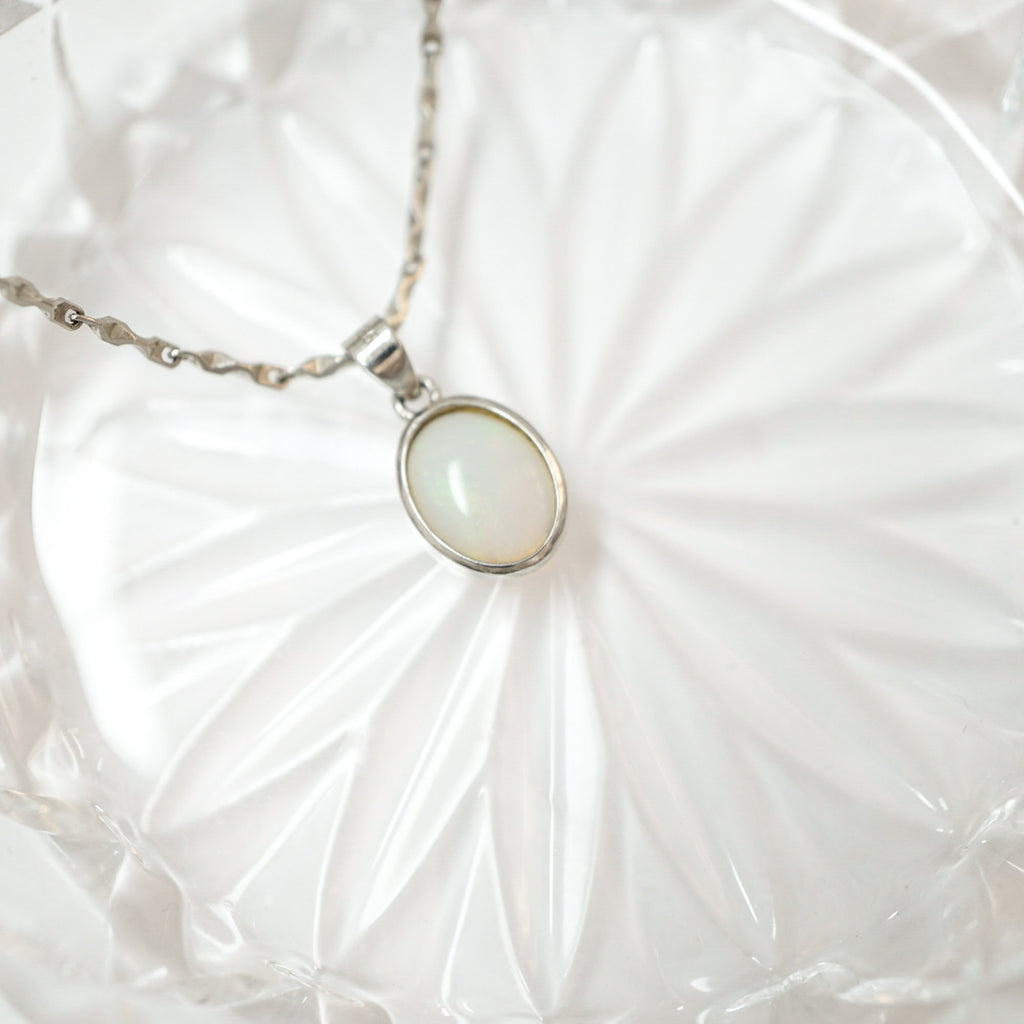 A silver chain necklace with an oval white iridescent pendant, resting on a white textured glass plate.