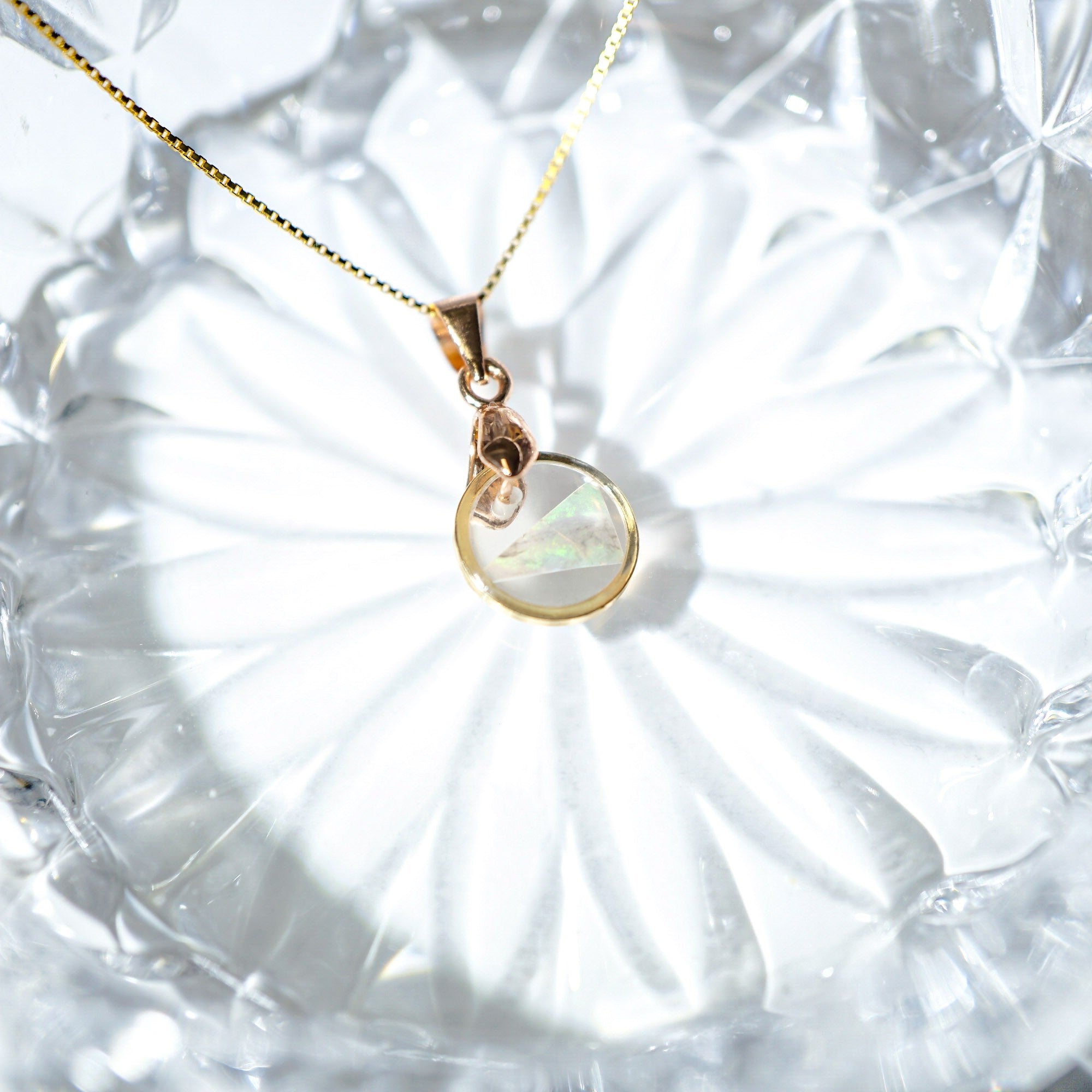 A gold chain necklace with a pendant featuring a milky white iridescent stone and a small gold heart-shaped accent, displayed on a textured clear glass surface.