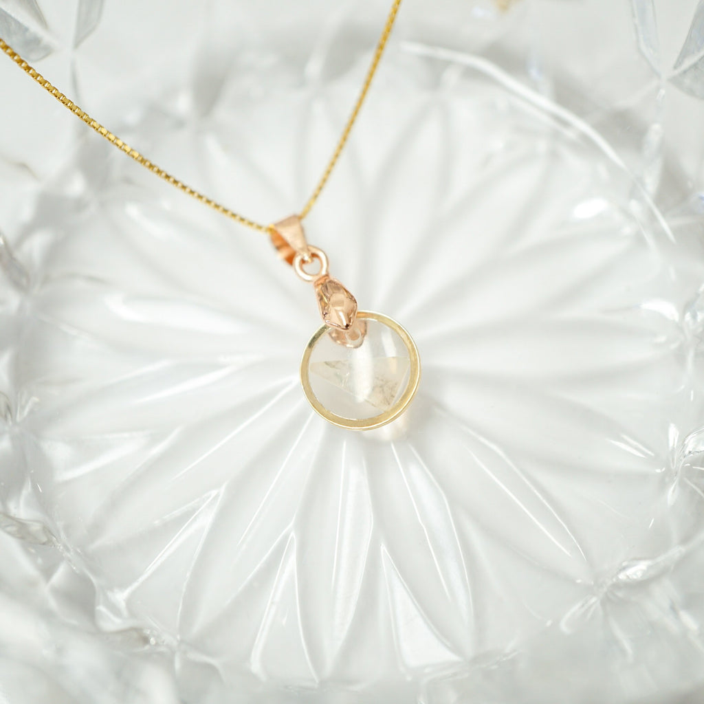 A delicate gold chain necklace with a rose gold pendant featuring a clear faceted gemstone, displayed on a textured white glass plate.
