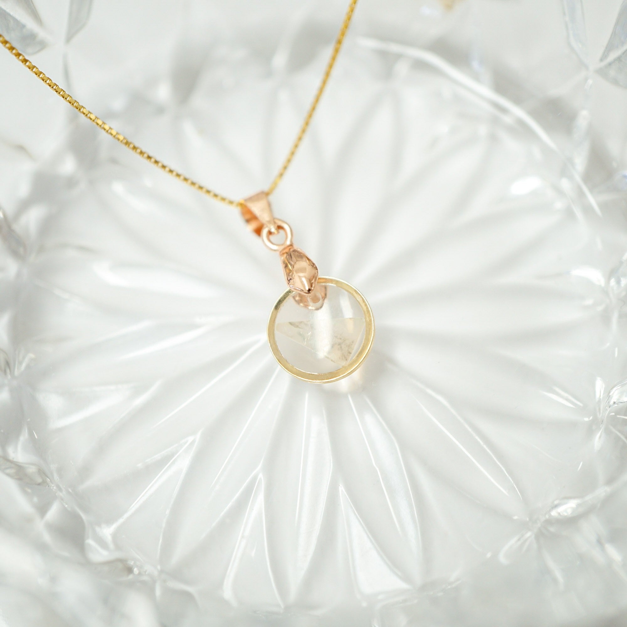 A delicate gold chain necklace with a rose gold pendant featuring a clear faceted gemstone, displayed on a textured white glass plate.