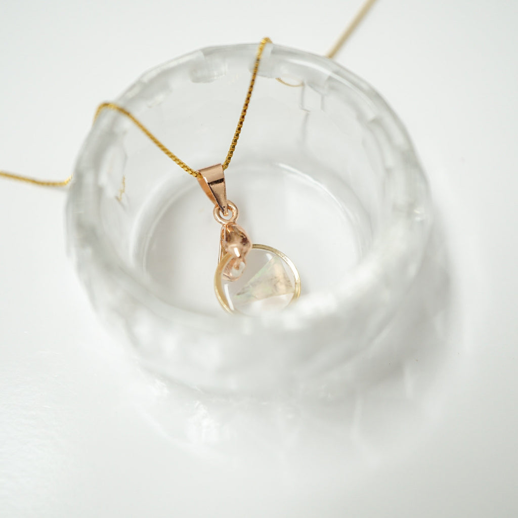 A delicate gold chain necklace with a rose gold pendant featuring a clear, organic-shaped stone, displayed on a textured clear glass dish against a white background.