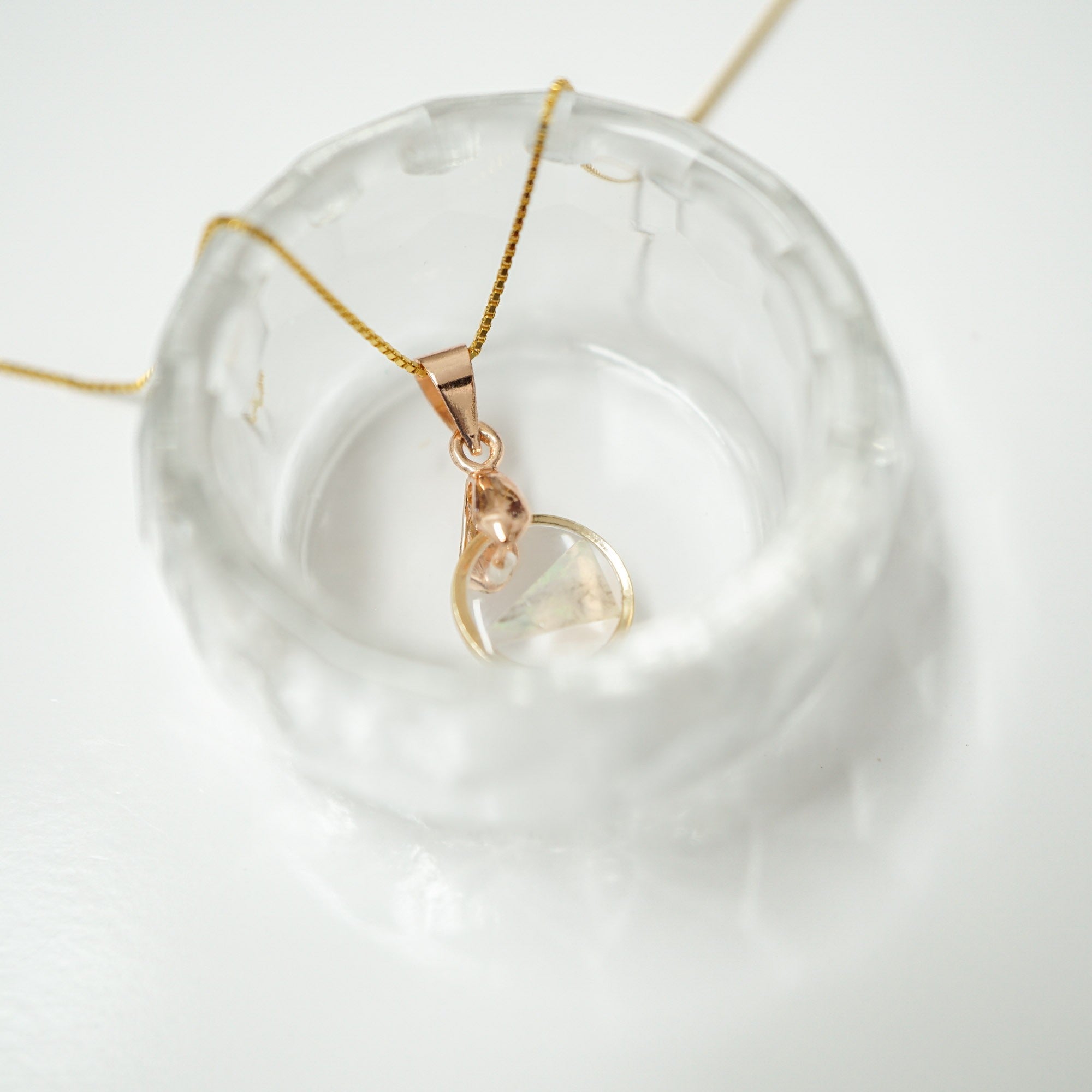 A delicate gold chain necklace with a rose gold pendant featuring a clear, organic-shaped stone, displayed on a textured clear glass dish against a white background.