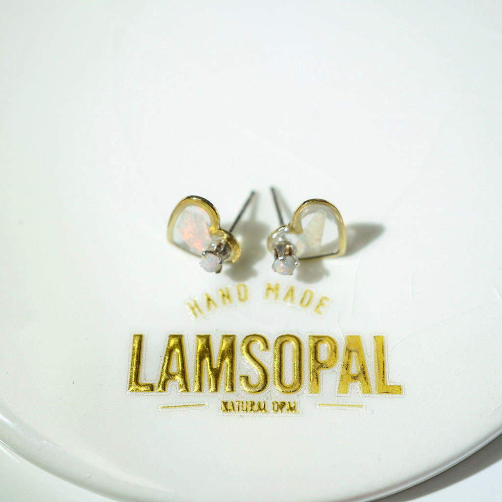 A pair of gold heart-shaped stud earrings with white opal inlays and clear crystal accents, displayed on a white circular surface with gold 'LAMSOPAL' branding.