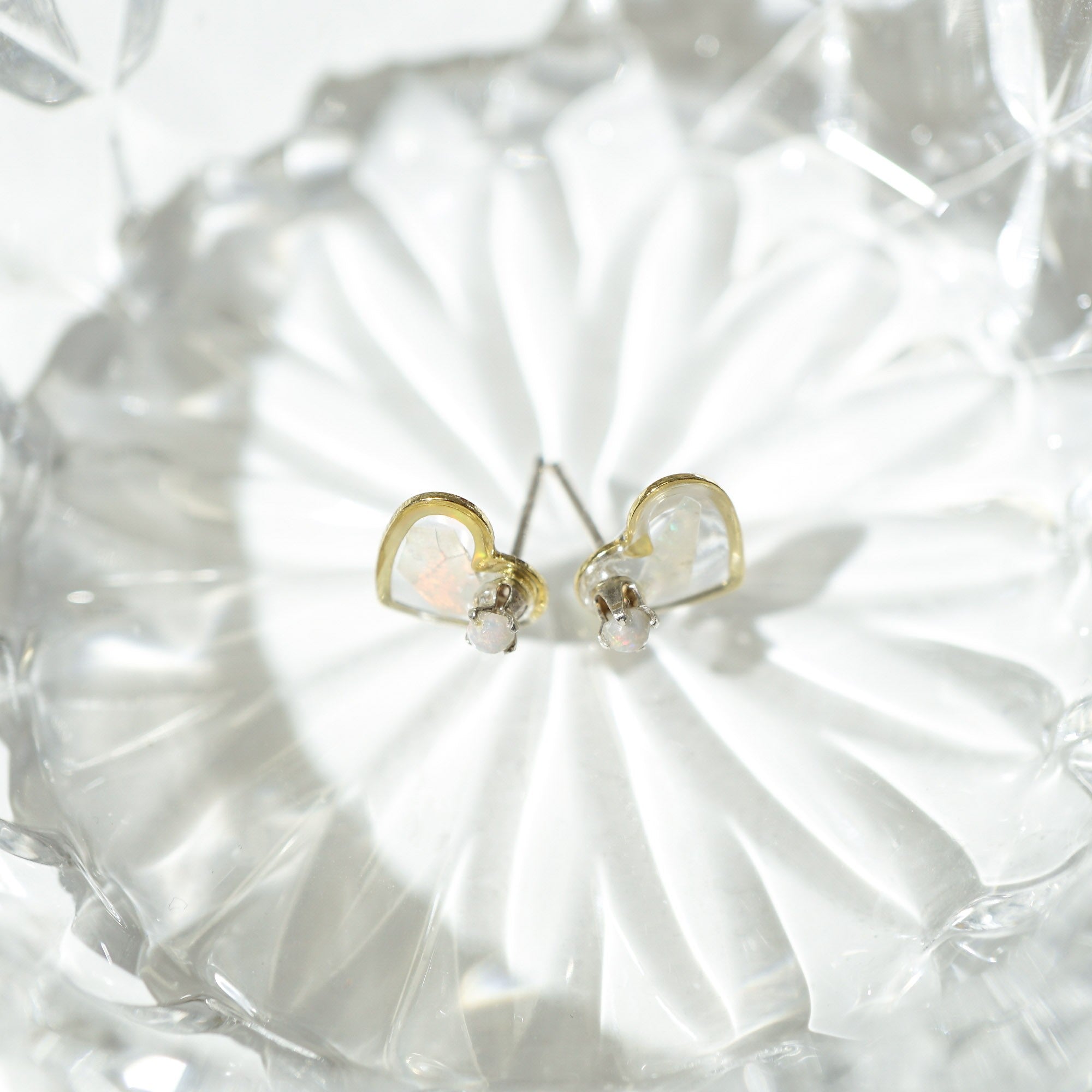 A pair of gold heart-shaped stud earrings with iridescent white opal inlays, displayed on a clear glass dish.