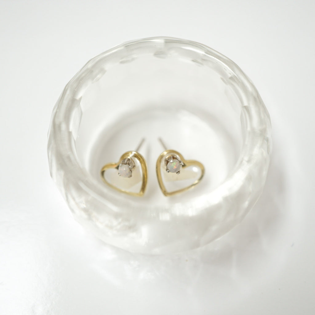 A pair of small gold heart stud earrings with clear oval gemstones, displayed inside a clear textured glass bowl on a white background.