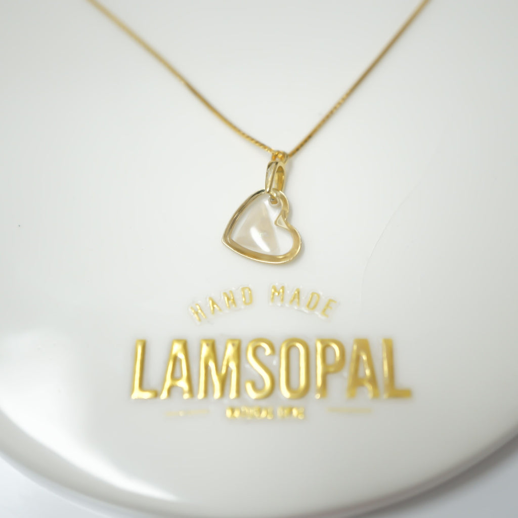 A gold heart-shaped pendant necklace with a clear glass or acrylic center, displayed on a white circular stand engraved with the brand name 'LAMSOPAL'.