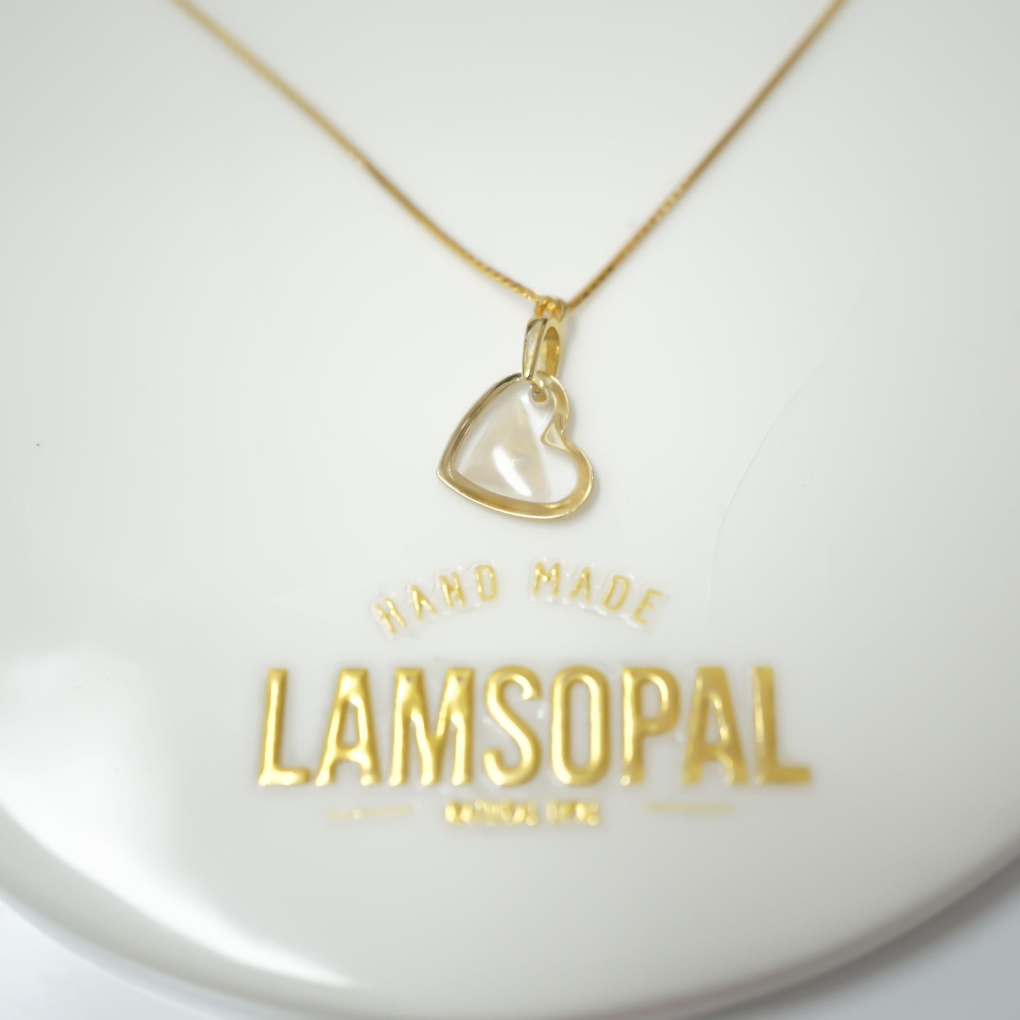A gold heart-shaped pendant necklace with a clear glass or acrylic center, displayed on a white circular stand engraved with the brand name 'LAMSOPAL'.