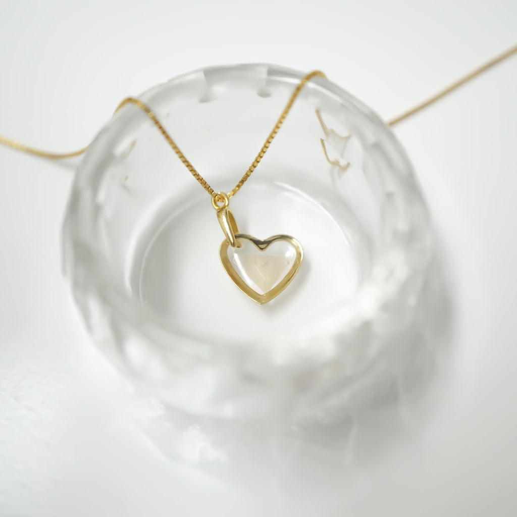 A gold heart-shaped pendant necklace with an open heart design, resting on a clear textured glass dish against a white background.