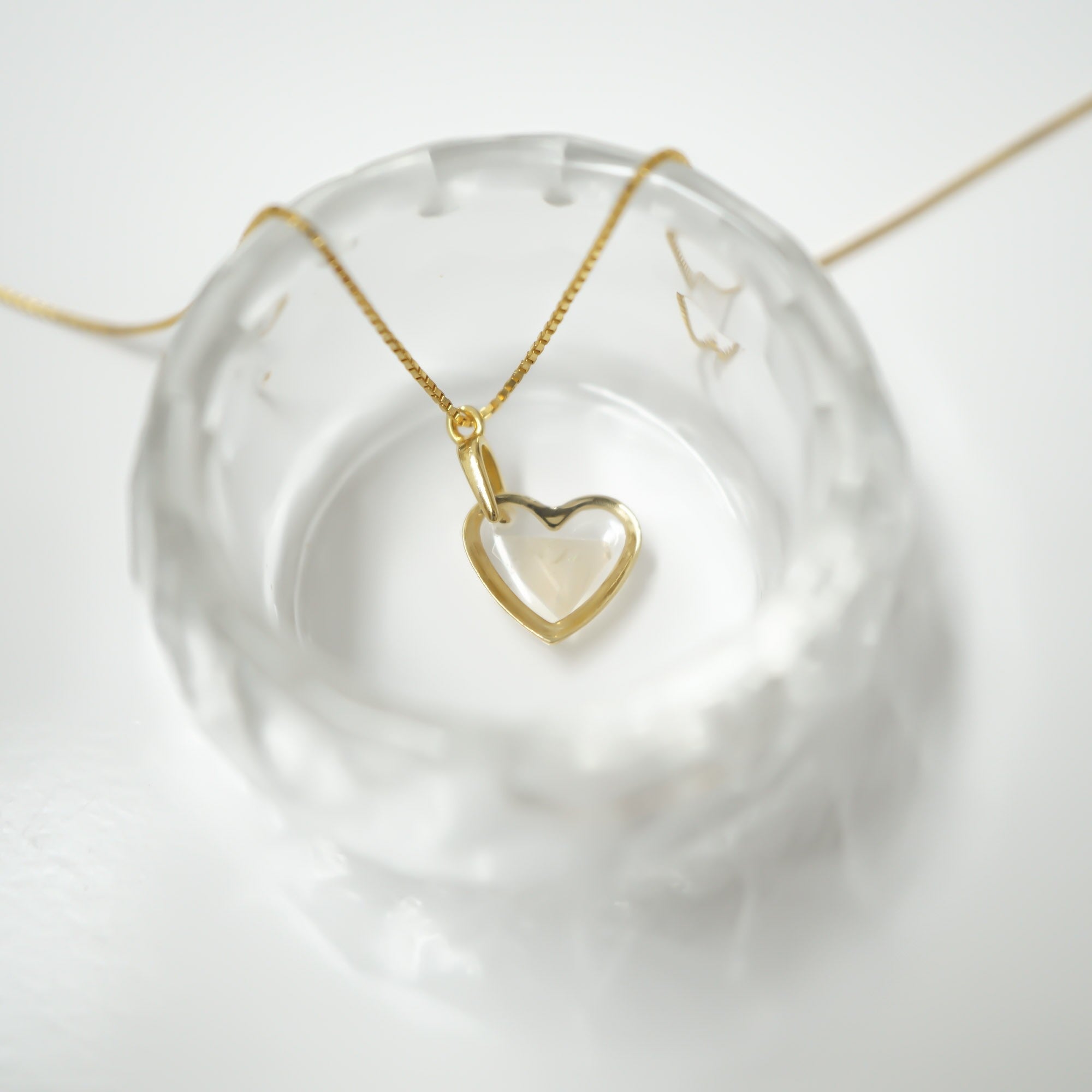 A gold heart-shaped pendant necklace with an open heart design, resting on a clear textured glass dish against a white background.