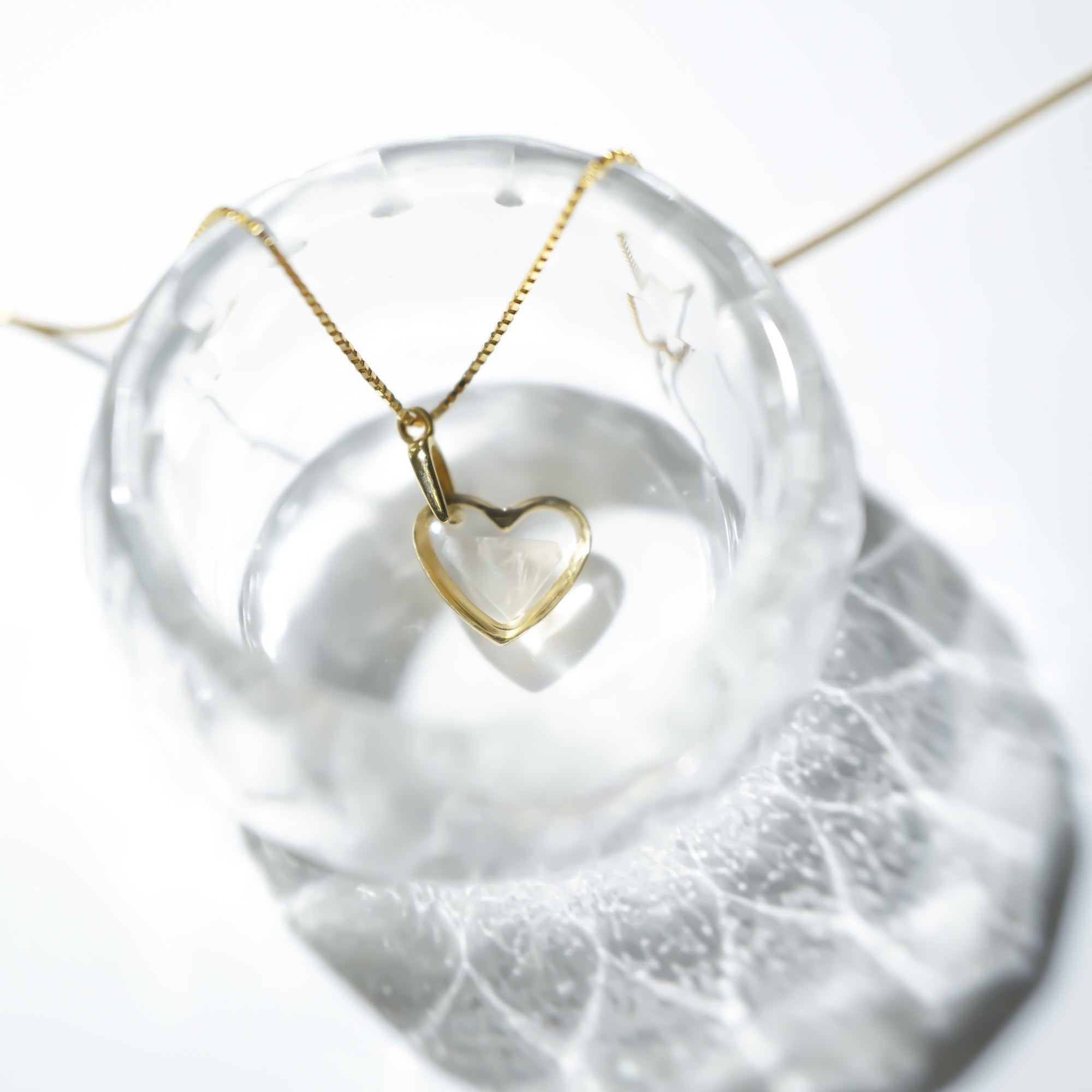 A delicate gold heart pendant necklace on a fine chain, displayed inside a clear glass bowl against a bright white background.