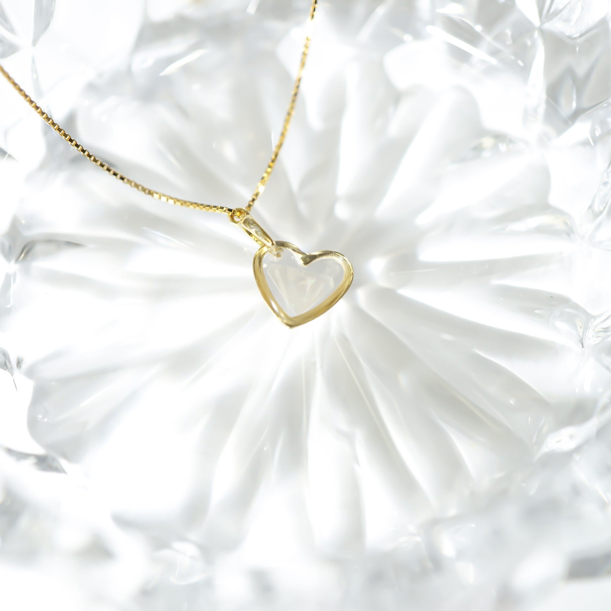 A delicate gold chain necklace with a thin, open heart-shaped pendant, displayed on a textured white glass plate.