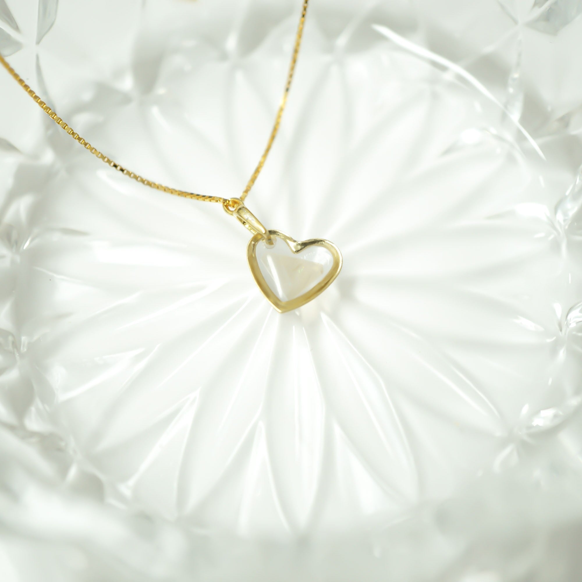 A delicate gold chain necklace with a heart-shaped pendant featuring a white inlay, displayed on a textured white surface.