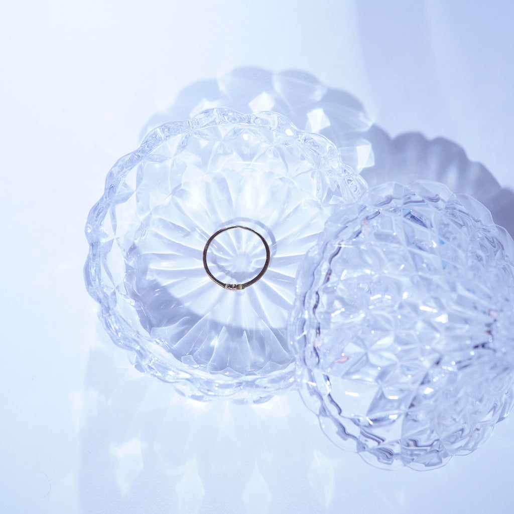 A thin silver ring displayed inside a clear, faceted glass dish with a matching lid, set against a soft, light blue background.