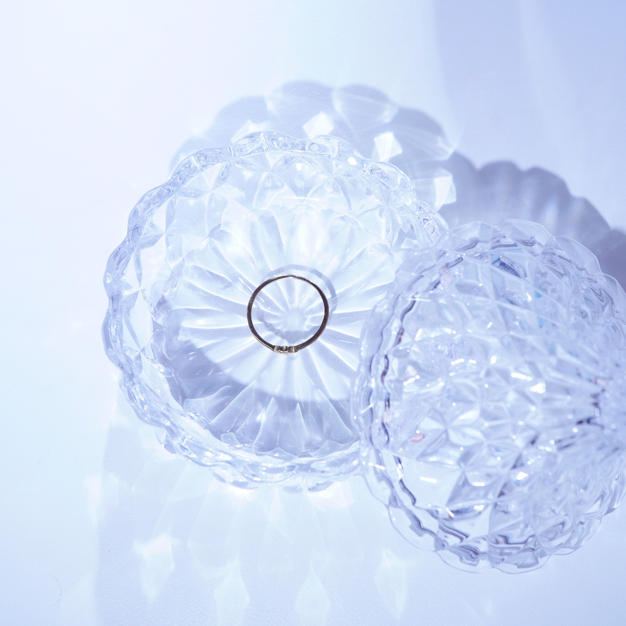 A thin silver ring displayed inside a clear, faceted glass dish with a matching lid, set against a soft, light blue background.