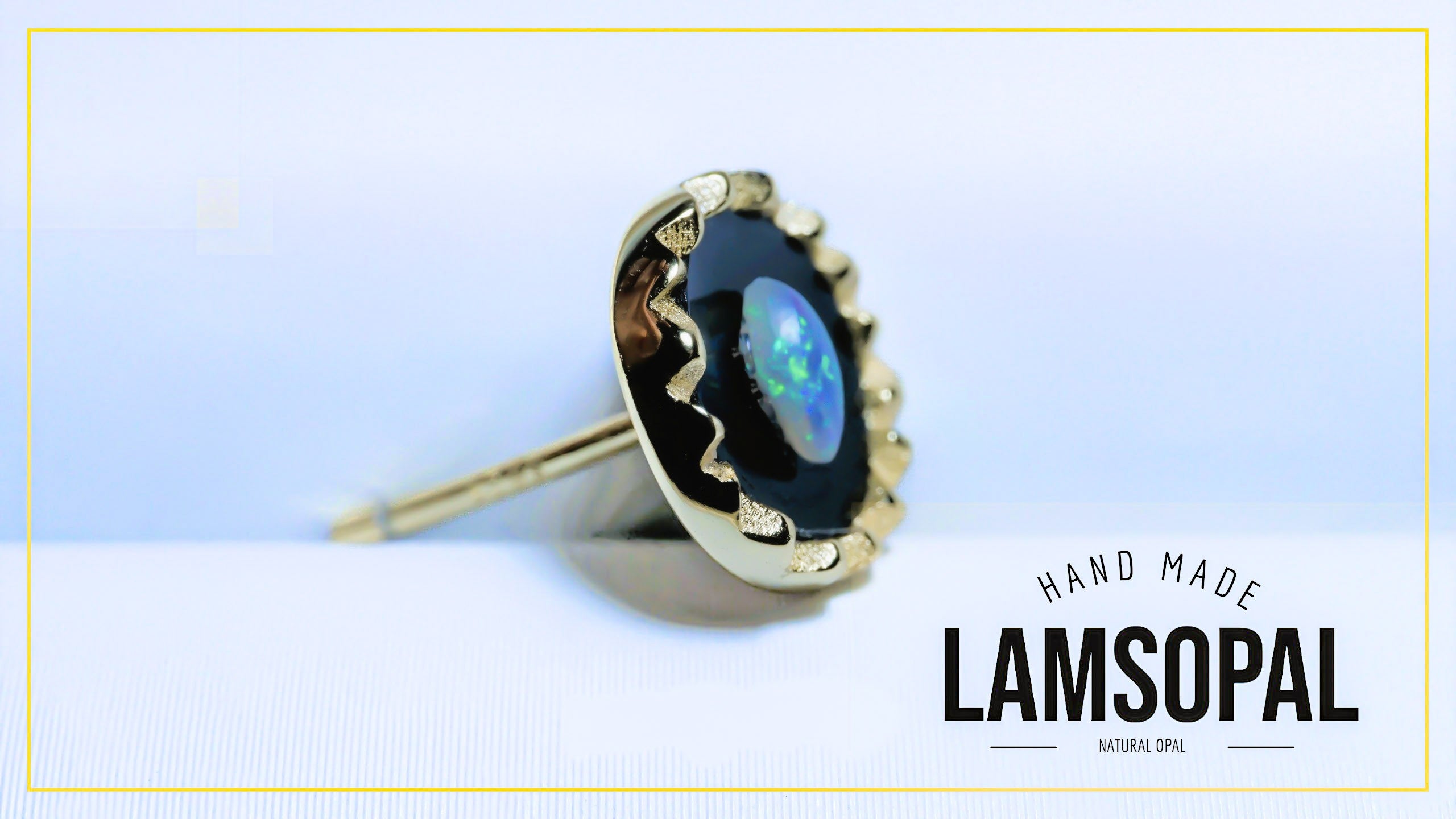 A single Lamsopal stud earring featuring a natural opal gemstone with iridescent blue and green play-of-color, set in a scalloped gold-tone bezel with a post backing. The earring is displayed on a light blue and white background with a gold border, and the text 'HAND MADE LAMSOPAL' is visible in the lower right.