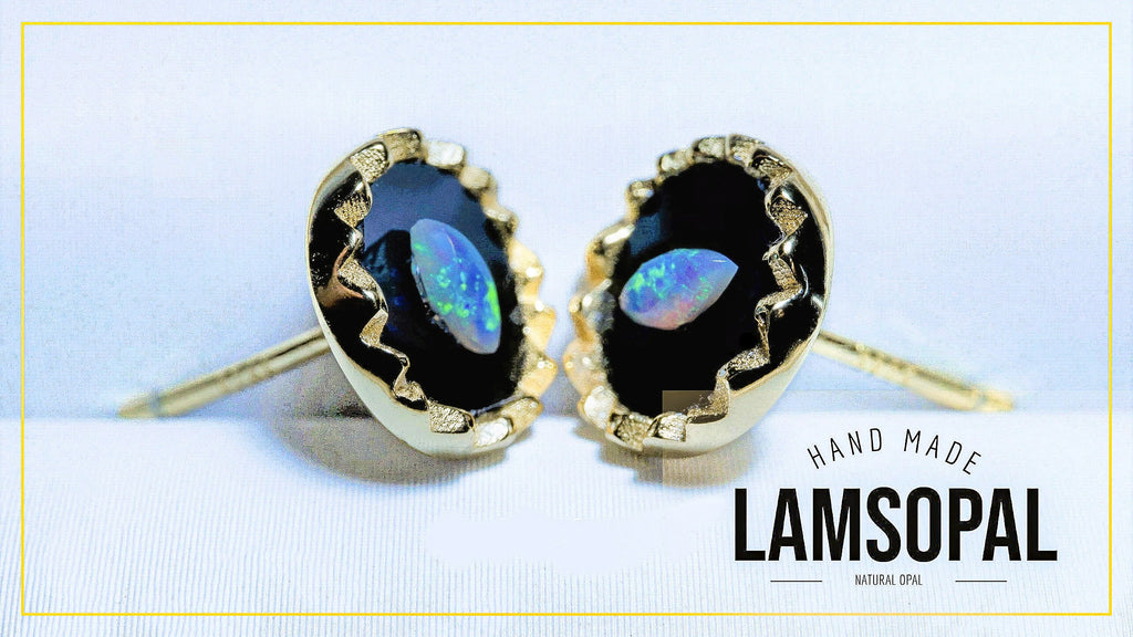 A pair of gold stud earrings with teardrop-shaped black opal gemstones showing blue and green flashes, set in a textured gold bezel. The image is set against a light blue background with a gold border, and features a text overlay on the bottom right that reads 'HAND MADE LAMSOPAL NATURAL OPAL'.