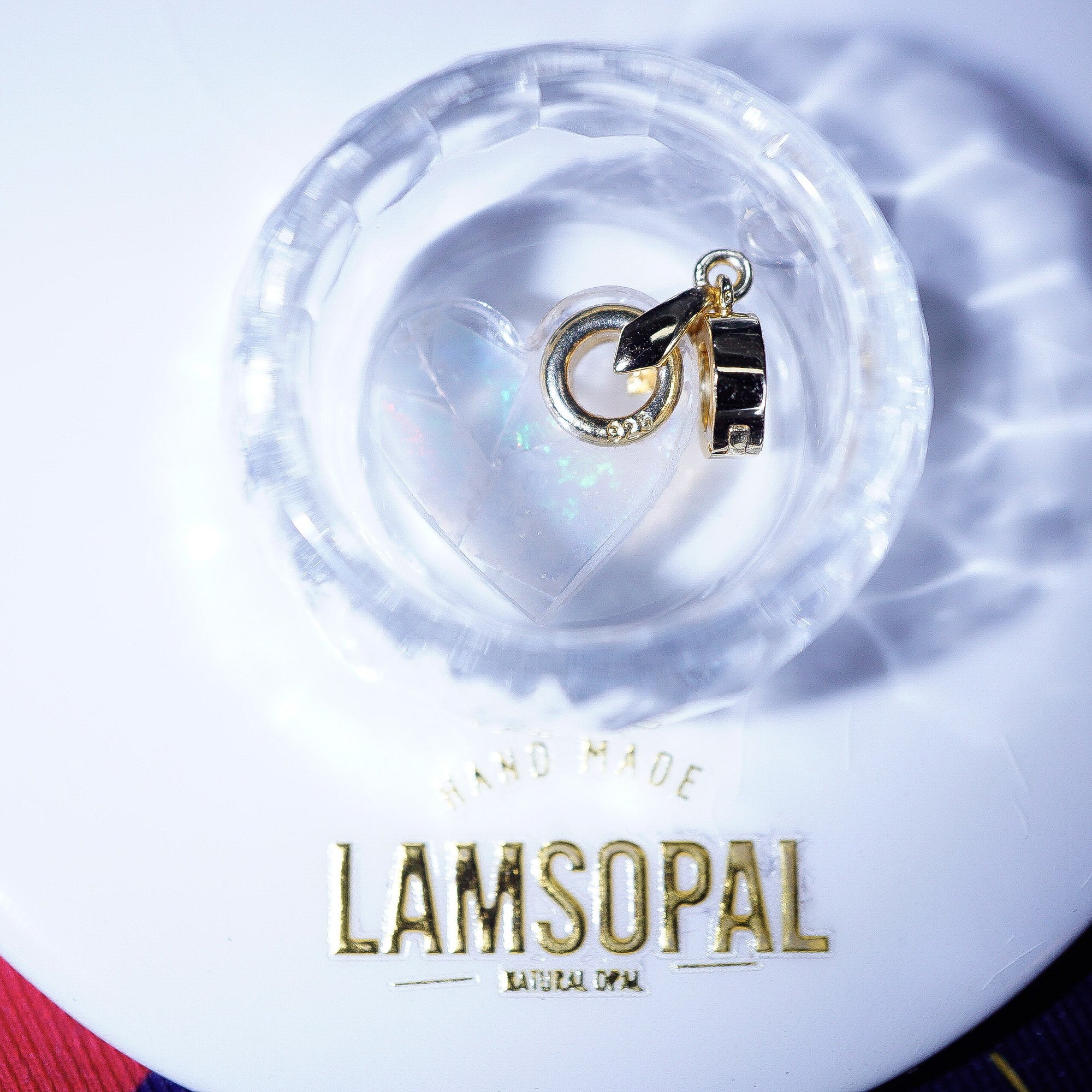 A gold and black enamel charm with a small heart and crescent moon design, resting inside a clear glass bowl on a white surface with the brand name 'LAMSOPAL' printed below.