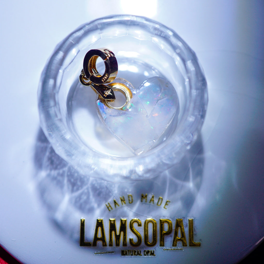 A handmade Lamsopal natural opal heart charm with a gold clasp, displayed inside a clear glass bowl on a white surface with a branded gold logo at the bottom.