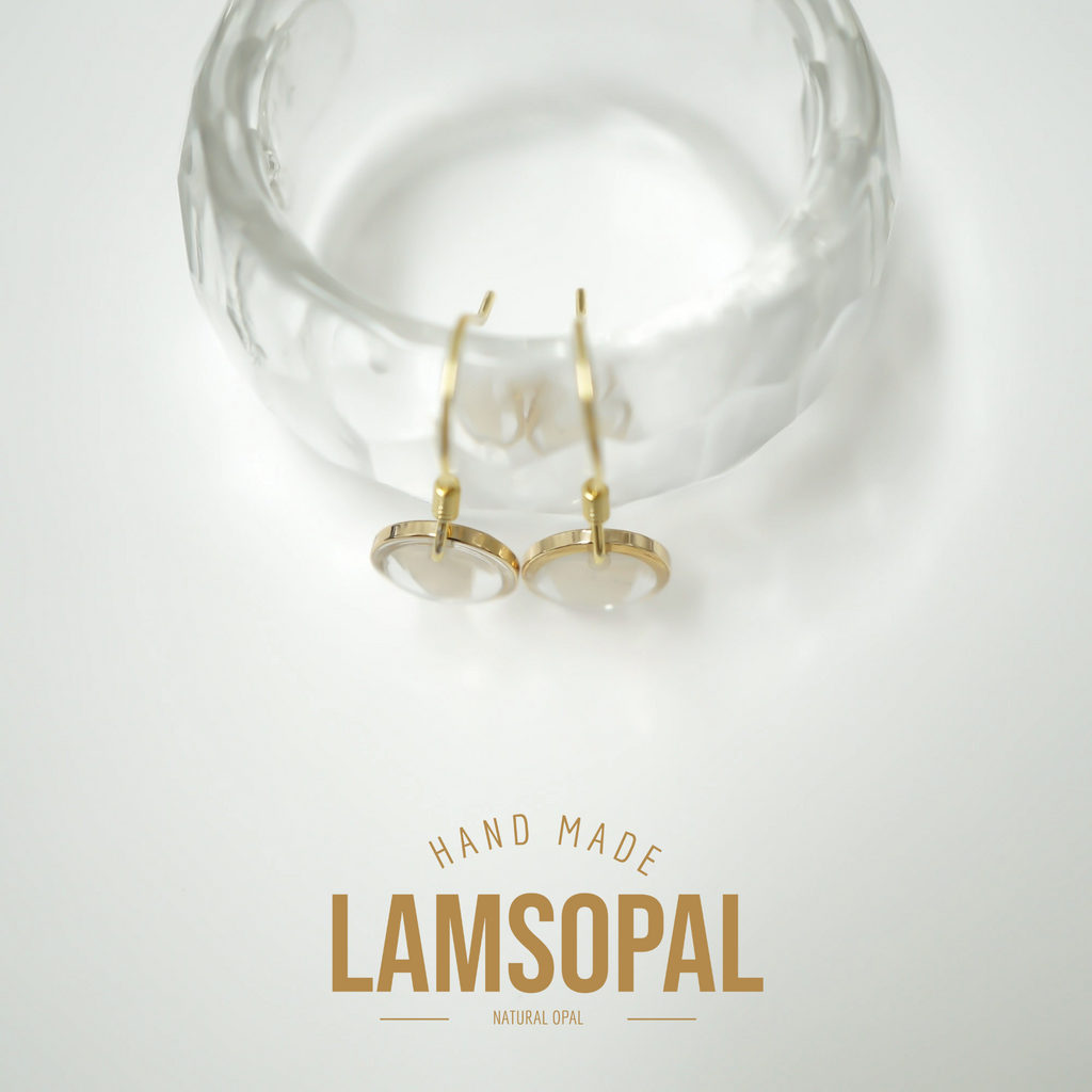 A pair of gold hoop earrings with oval clear stone settings, displayed on a textured clear glass stand against a white background, with the brand name 'LAMSOPAL' and 'HAND MADE NATURAL OPAL' printed below.
