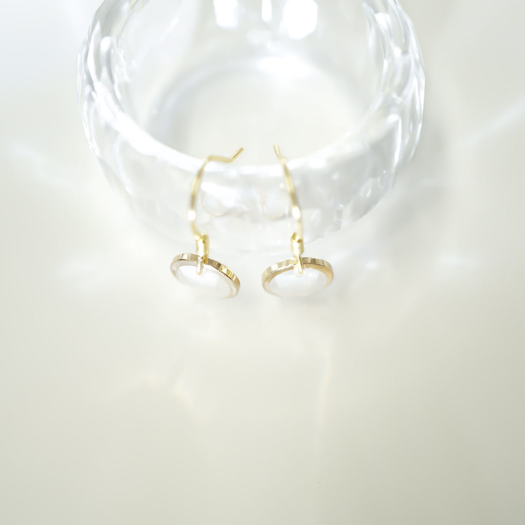 A pair of gold hoop earrings with oval white stone settings, displayed on a clear glass stand against a bright white background.