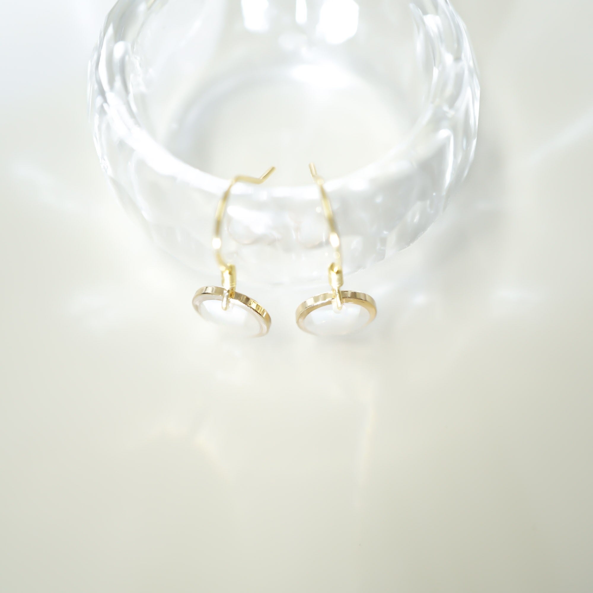 A pair of gold hoop earrings with oval white stone settings, displayed on a clear glass stand against a bright white background.
