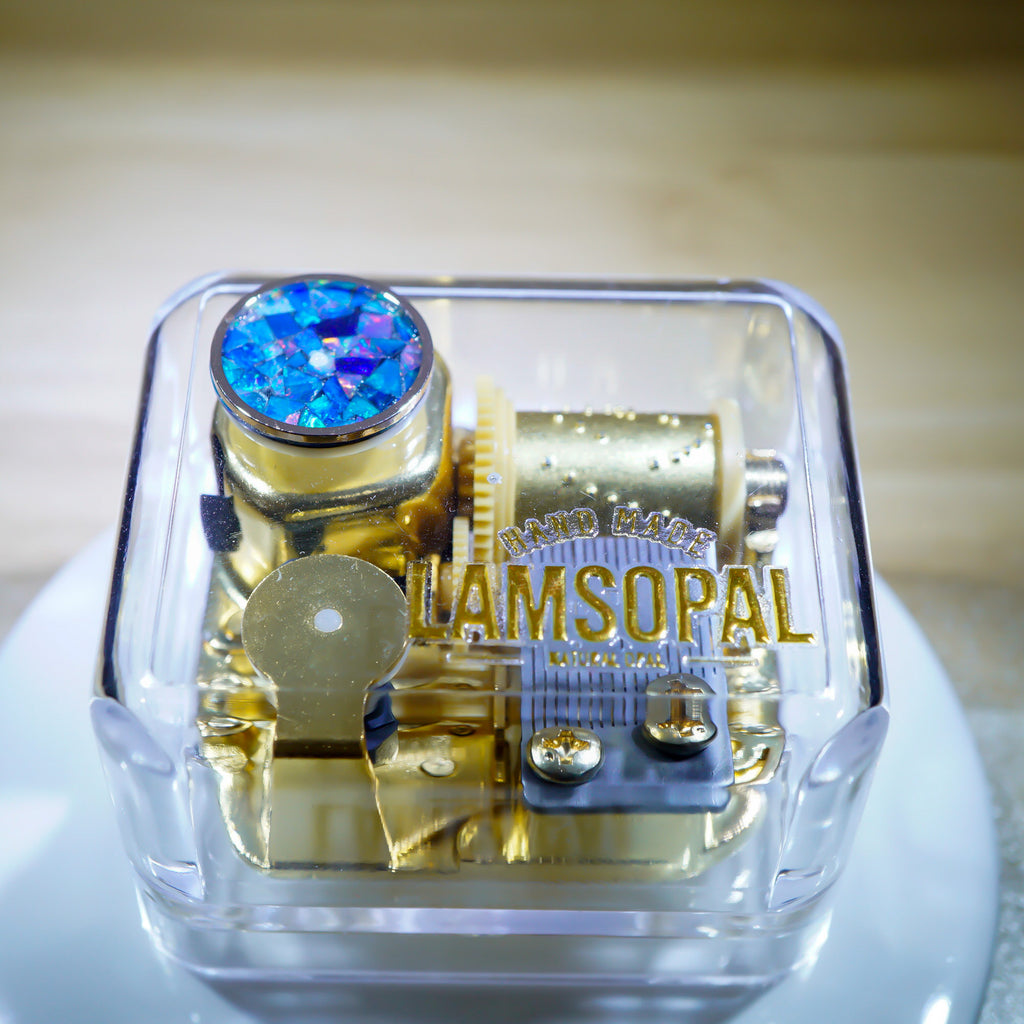 A transparent square jewelry box containing gold-toned mechanical gears and a blue iridescent rhinestone, with the brand name 'RAMSOPAL' printed in gold on the front.
