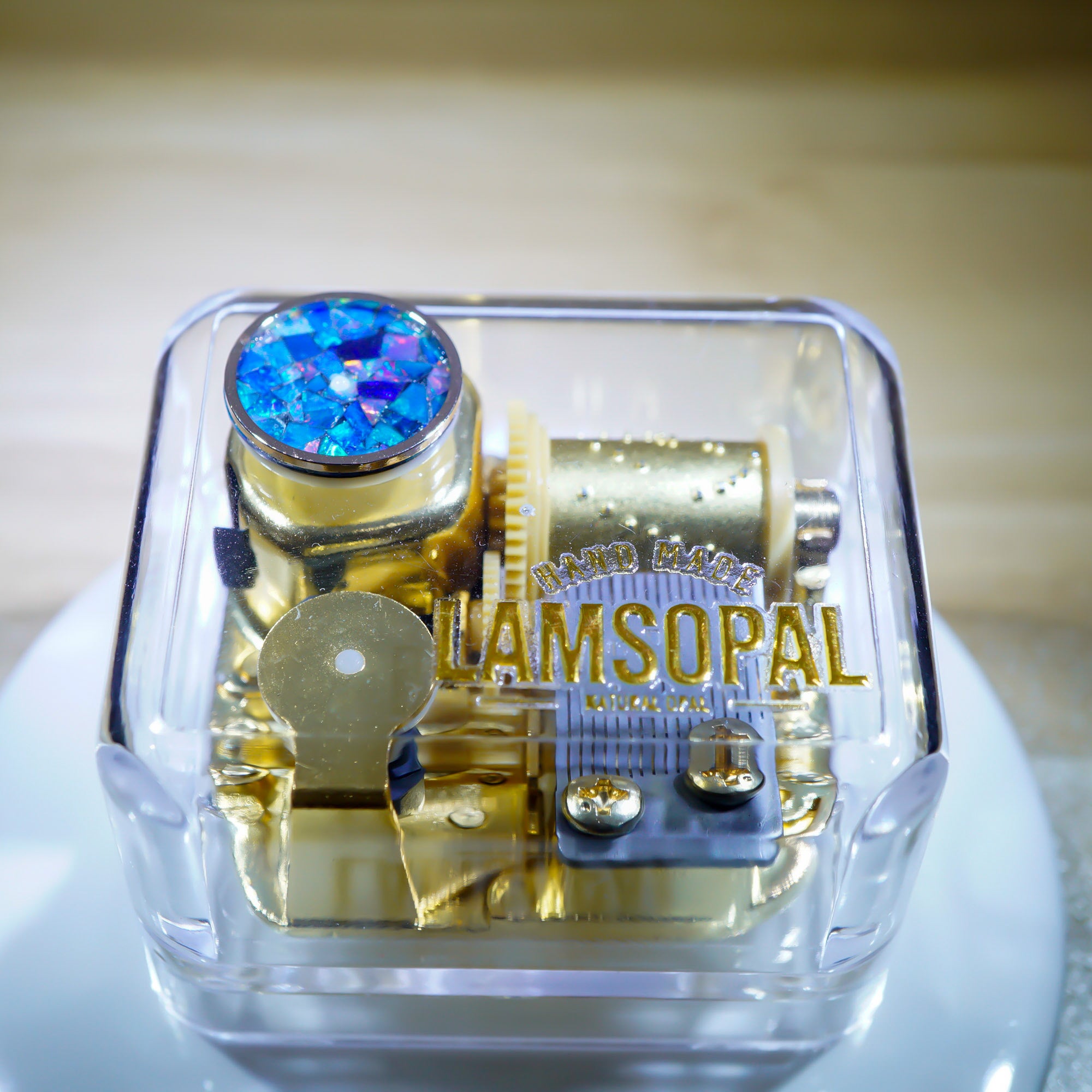 A transparent square jewelry box containing gold-toned mechanical gears and a blue iridescent rhinestone, with the brand name 'RAMSOPAL' printed in gold on the front.
