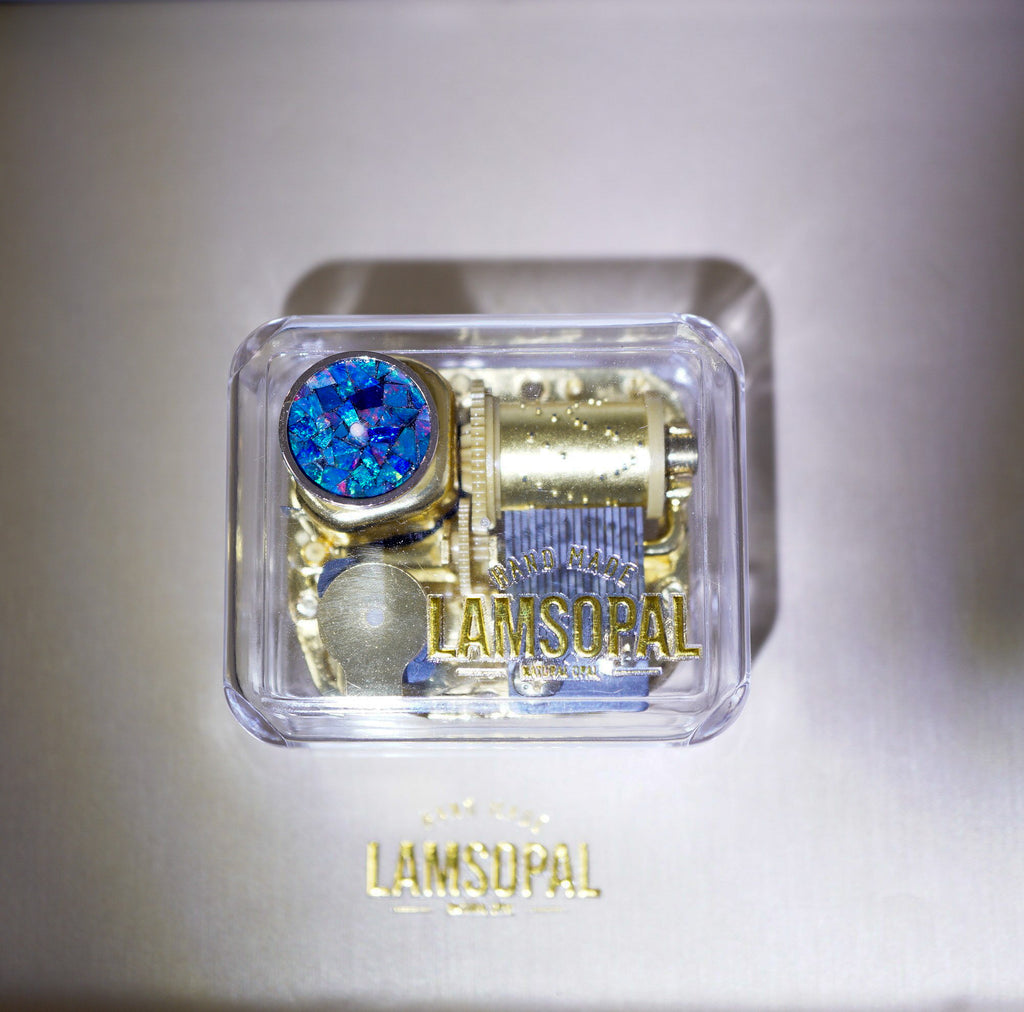 A Lamsopal luxury watch presentation box containing a gold-toned watch movement with a prominent blue iridescent round crystal and a blue striped component, set against a metallic silver background with the Lamsopal logo below.