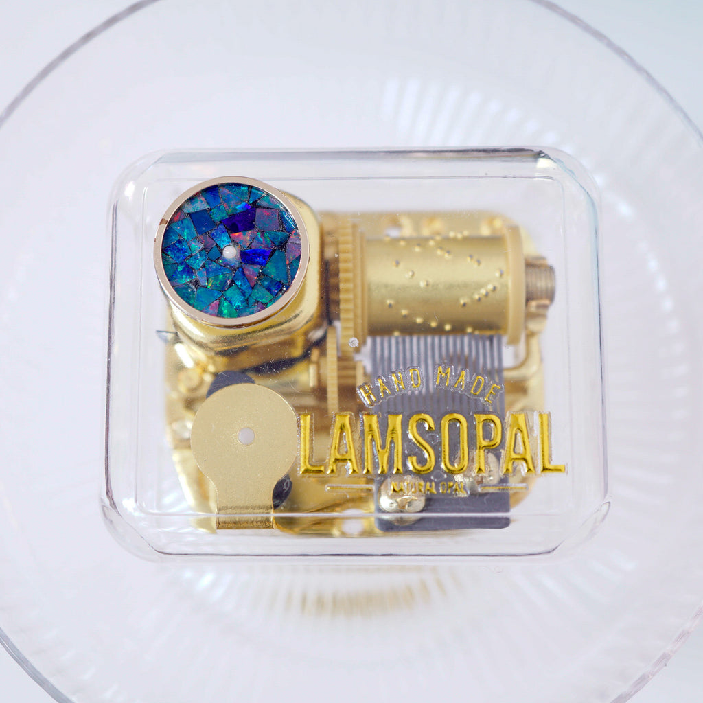 A gold-colored Lamsopal music box with a blue iridescent mosaic dial, sealed in a clear plastic protective case and placed on a white plate.