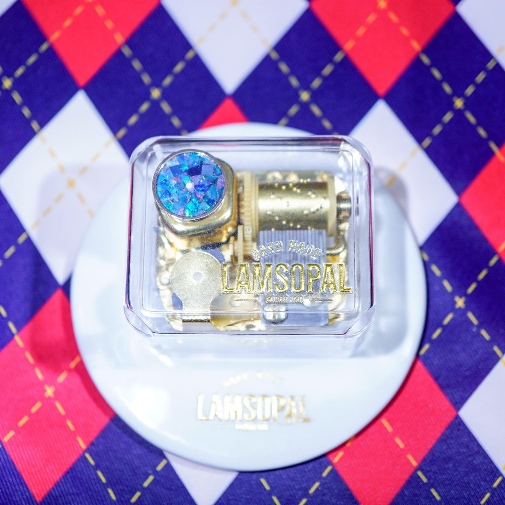 A gold-toned Lamsopal music box with a blue rhinestone, displayed in a clear plastic case on a white round stand over a red, white, and blue argyle patterned fabric background.