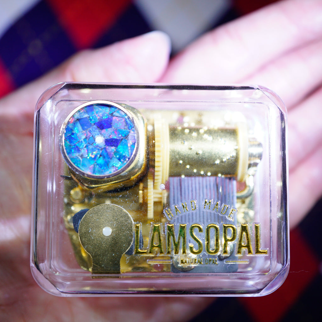 A hand holds a small clear plastic case containing a gold-toned mechanical music box with a blue iridescent stone and the text 'HAND MADE LAMSOPAL' printed on the front.
