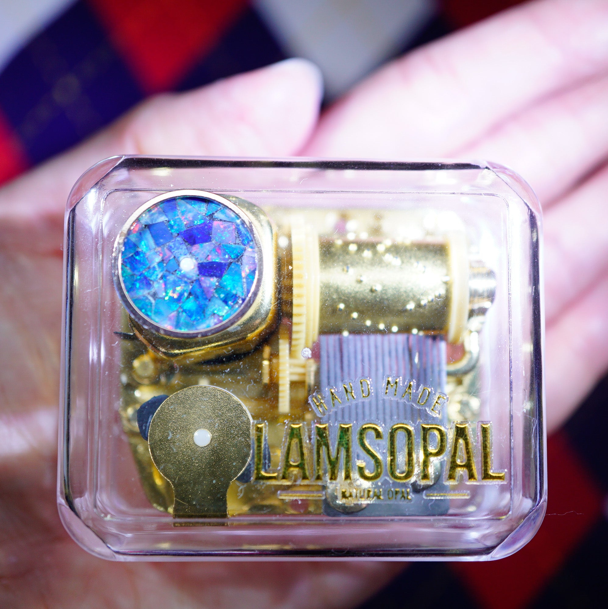 A hand holds a small clear plastic case containing a gold-toned mechanical music box with a blue iridescent stone and the text 'HAND MADE LAMSOPAL' printed on the front.
