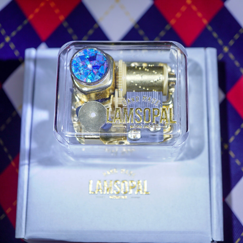 A gold-toned Lamsopal mechanical watch or toy mechanism displayed inside a clear plastic case on top of a branded light blue box, with a red, navy, and white argyle patterned surface in the background.