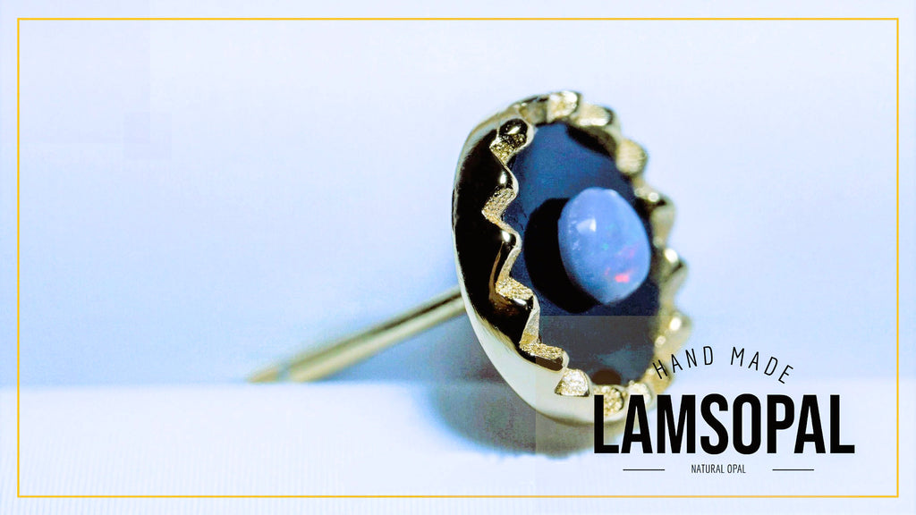 A gold ring featuring a large, irregularly shaped dark blue opal with a smooth light blue center, set in a textured gold bezel. The ring is displayed on a light blue and white background with a gold border, and a text overlay reads 'HAND MADE LAMSOPAL'.