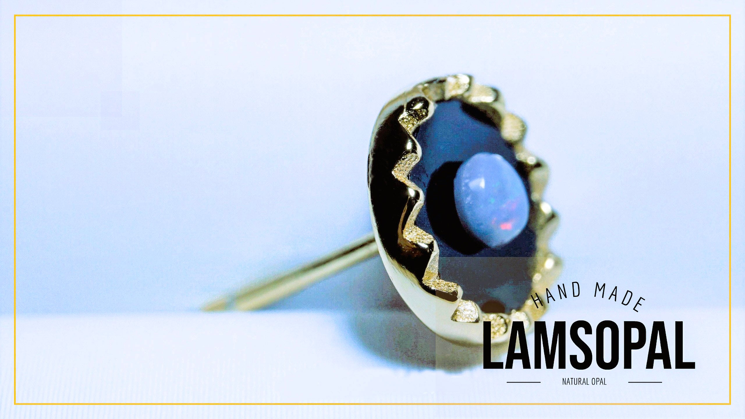 A gold ring featuring a large, irregularly shaped dark blue opal with a smooth light blue center, set in a textured gold bezel. The ring is displayed on a light blue and white background with a gold border, and a text overlay reads 'HAND MADE LAMSOPAL'.