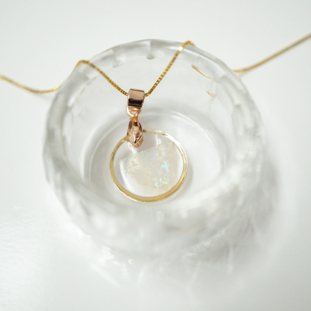 A delicate gold chain necklace with a rose gold pendant featuring a round iridescent white stone, displayed on a textured clear glass dish against a white background.