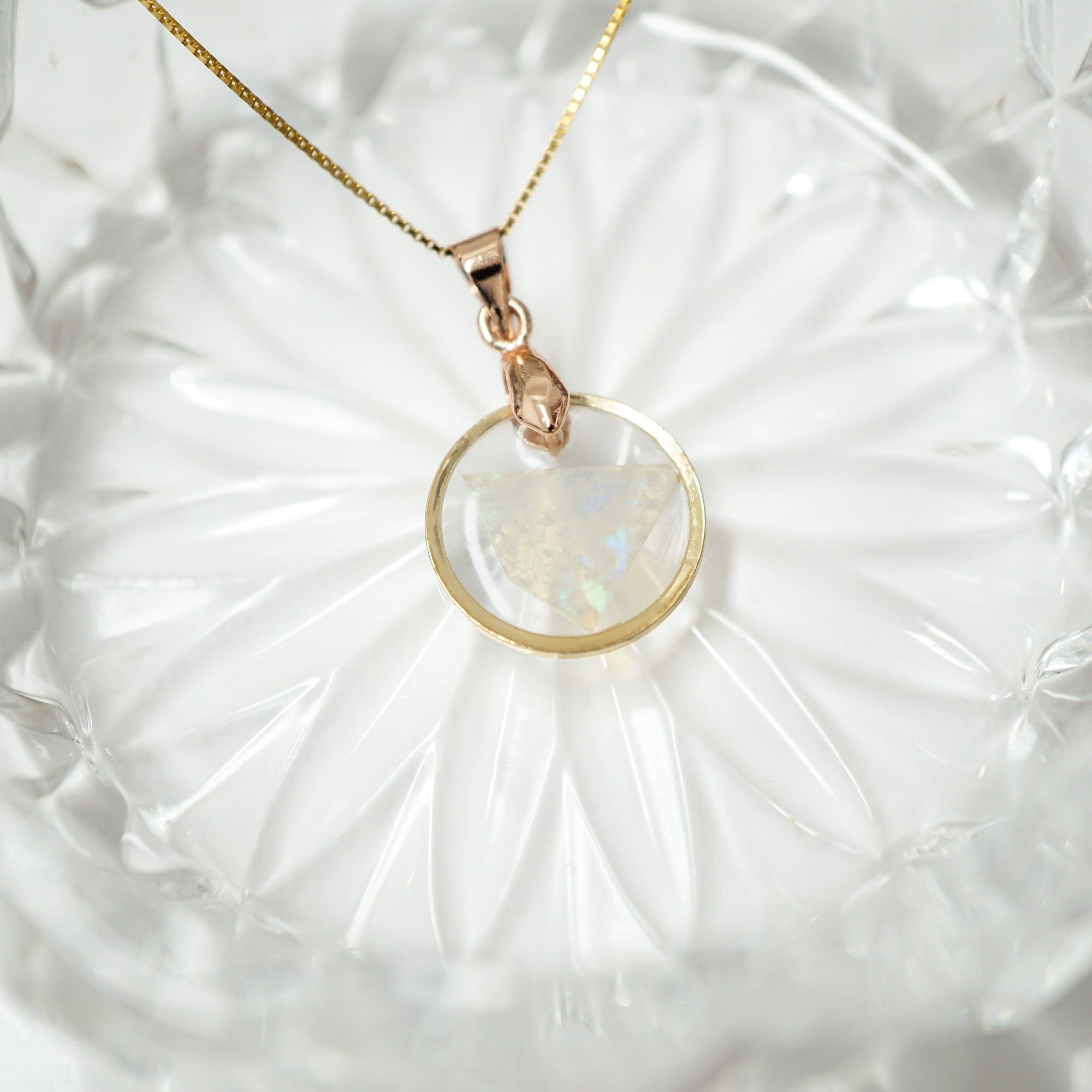 A gold chain necklace with a circular pendant featuring a milky white iridescent stone, displayed on a textured white glass dish.