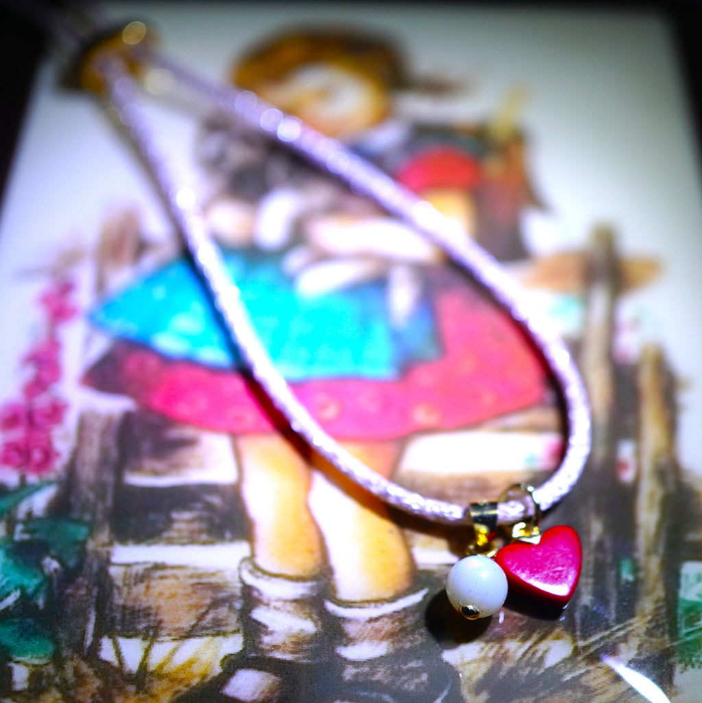 A silver-toned beaded necklace with a dangling charm featuring a bright pink heart, a white bead, and gold-toned accents, resting on a colorful illustrated card.