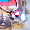 A close-up of a bracelet with a textured silver band and two charms: a white pearl bead and a shiny pink heart charm, resting on a printed mushroom illustration.