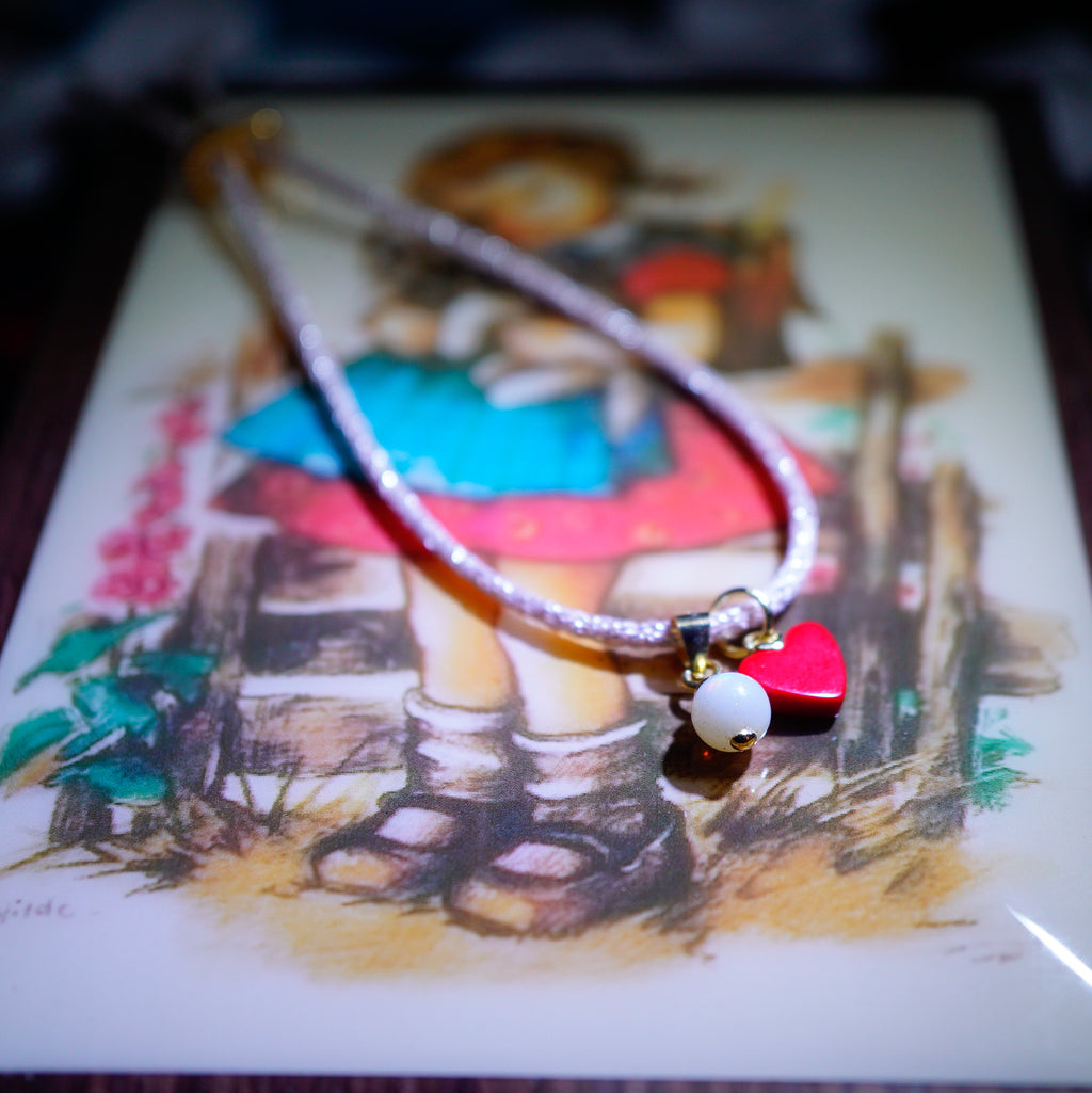 A beaded necklace with a silver-toned cord, featuring a red heart-shaped charm, a white pearl bead, and a small gold bell, displayed on top of a vintage-style illustrated card.