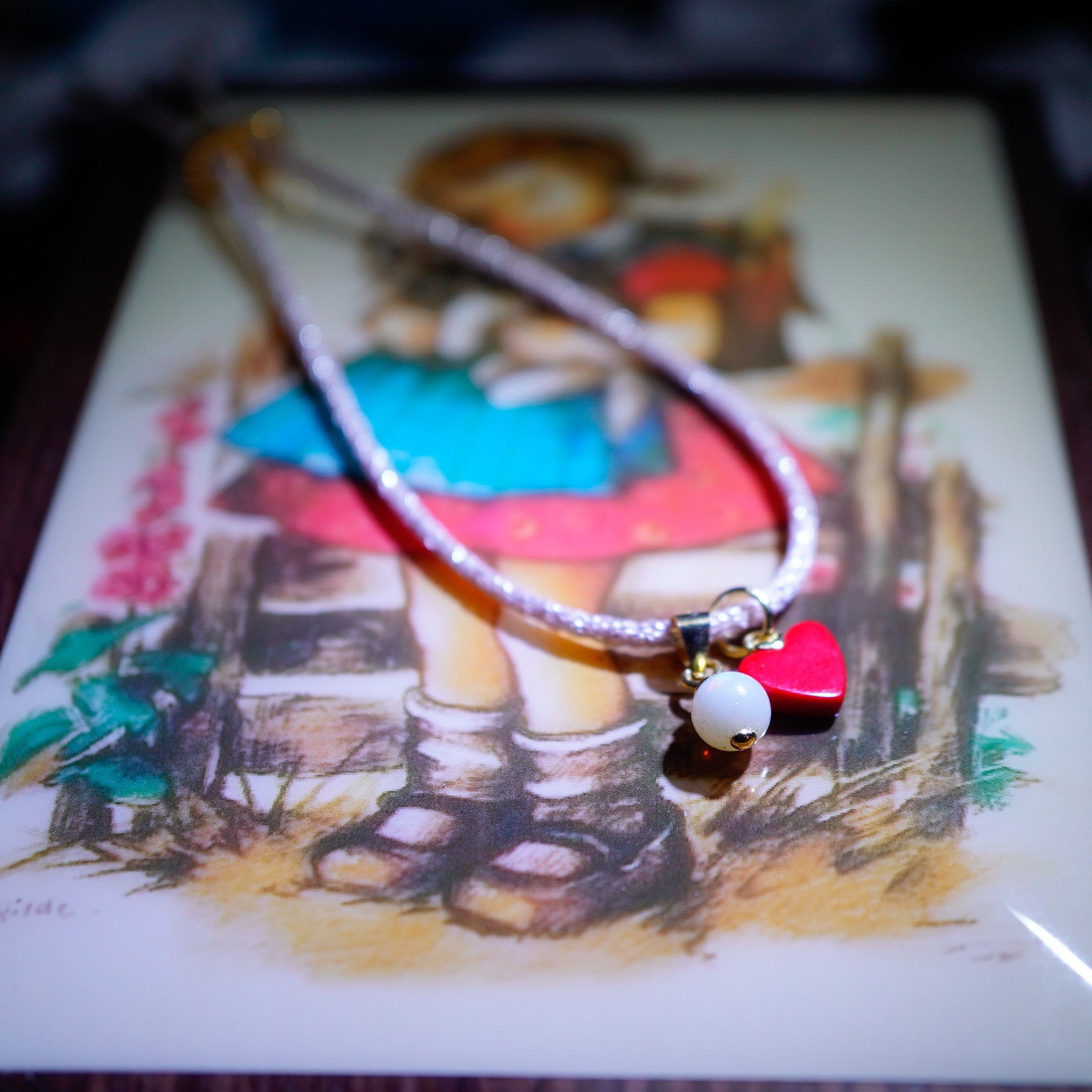 A beaded necklace with a silver-toned cord, featuring a red heart-shaped charm, a white pearl bead, and a small gold bell, displayed on top of a vintage-style illustrated card.