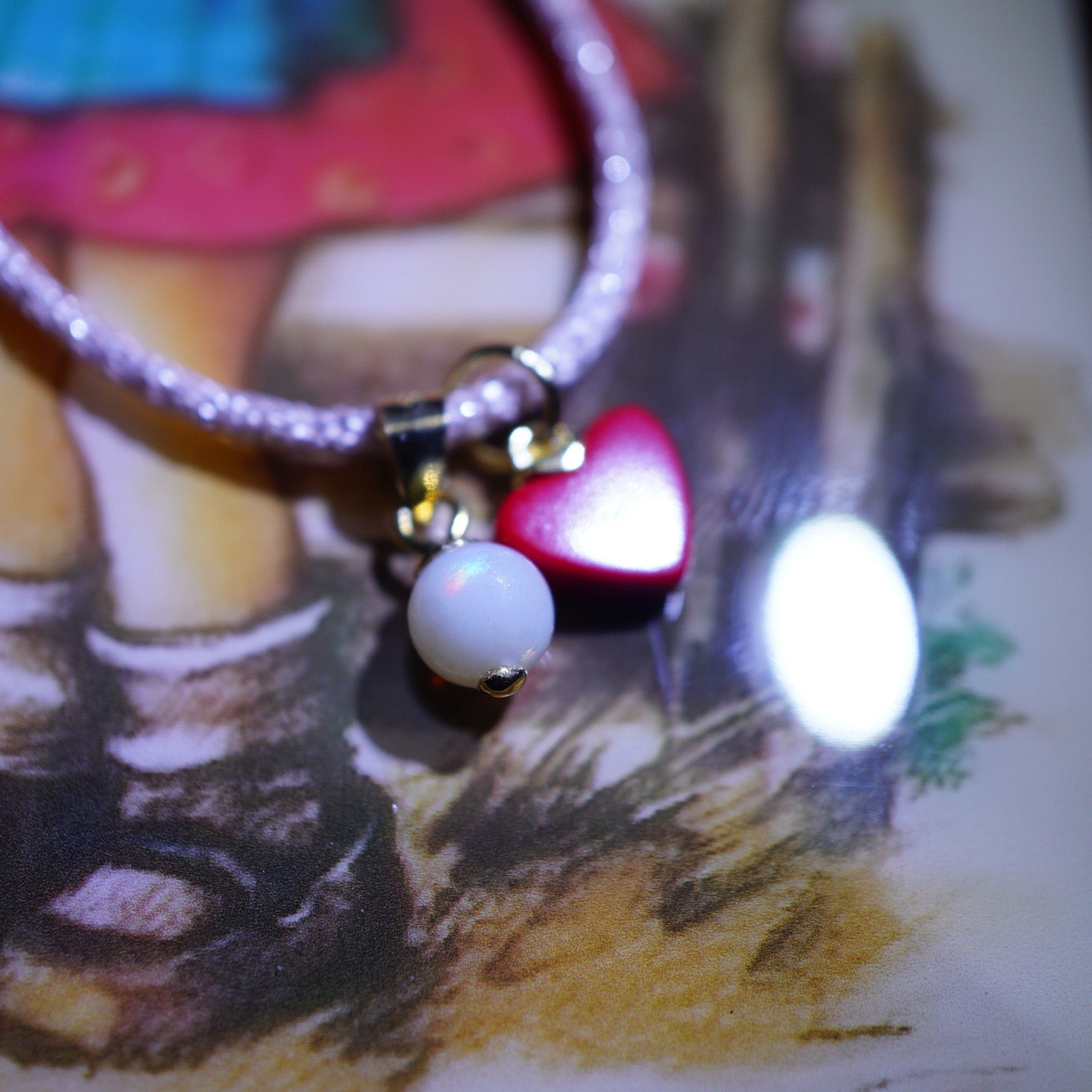 A close-up of a bracelet with a silver-toned beaded band, featuring a gold clasp holding a glossy red heart charm and a white pearl bead.