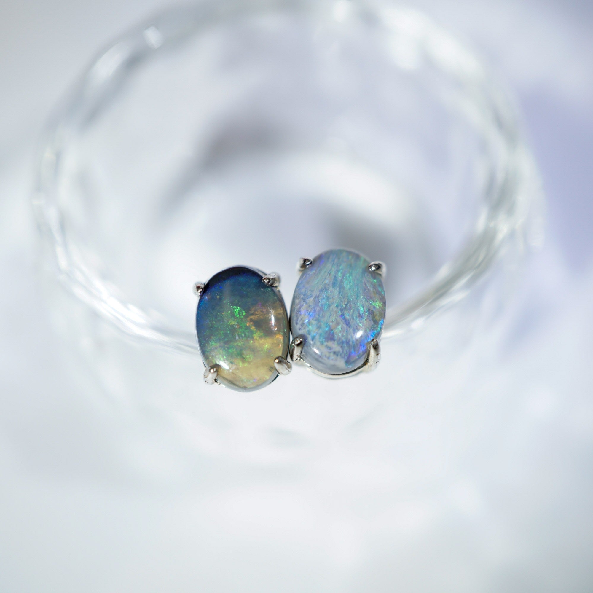 A pair of oval stud earrings with iridescent blue-green opal-like stones in silver settings, resting on a clear glass dish against a soft white background.