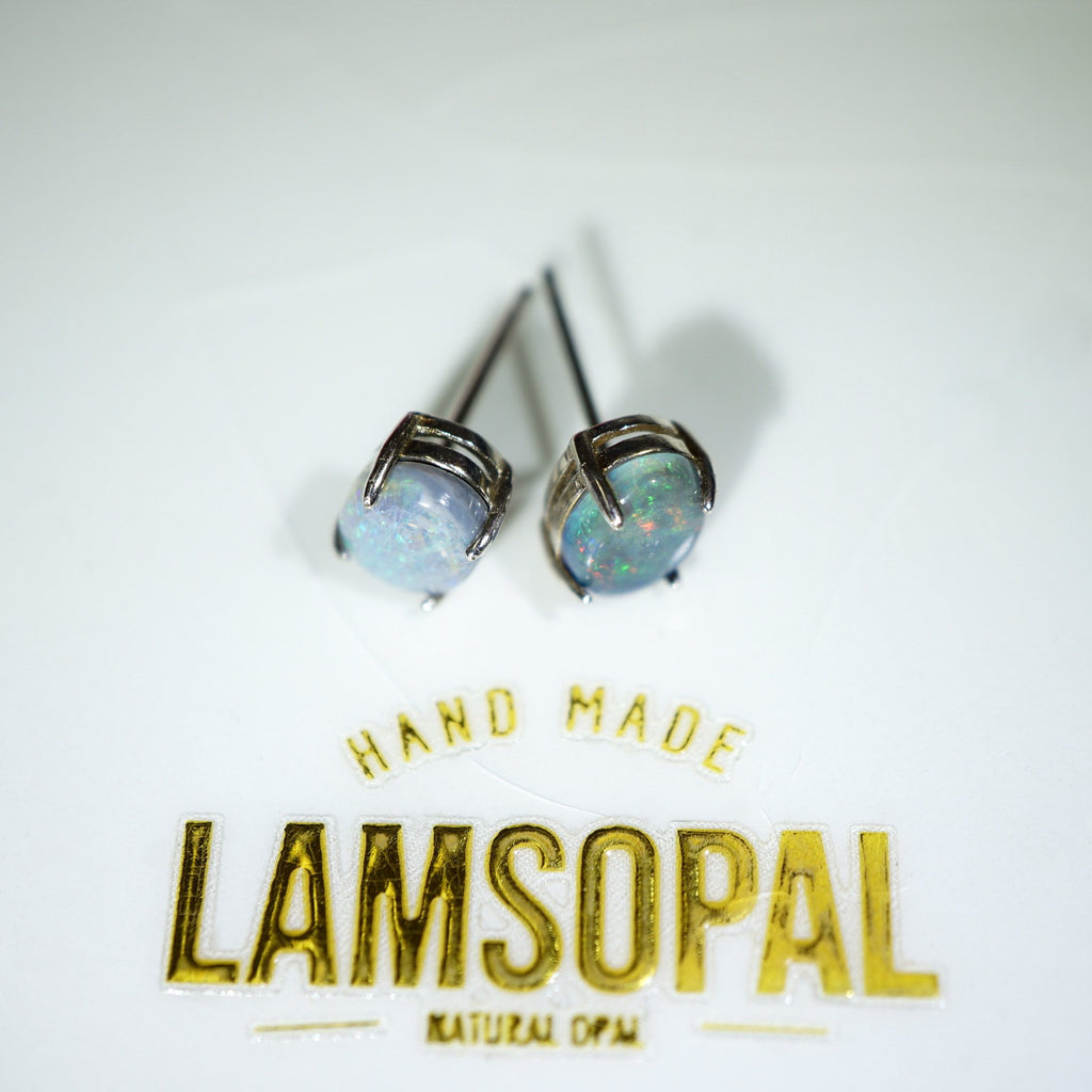 A pair of Lamsopal natural opal stud earrings with silver-toned settings, displayed on a white surface above gold foil text that reads 'HAND MADE LAMSOPAL NATURAL OPM'.