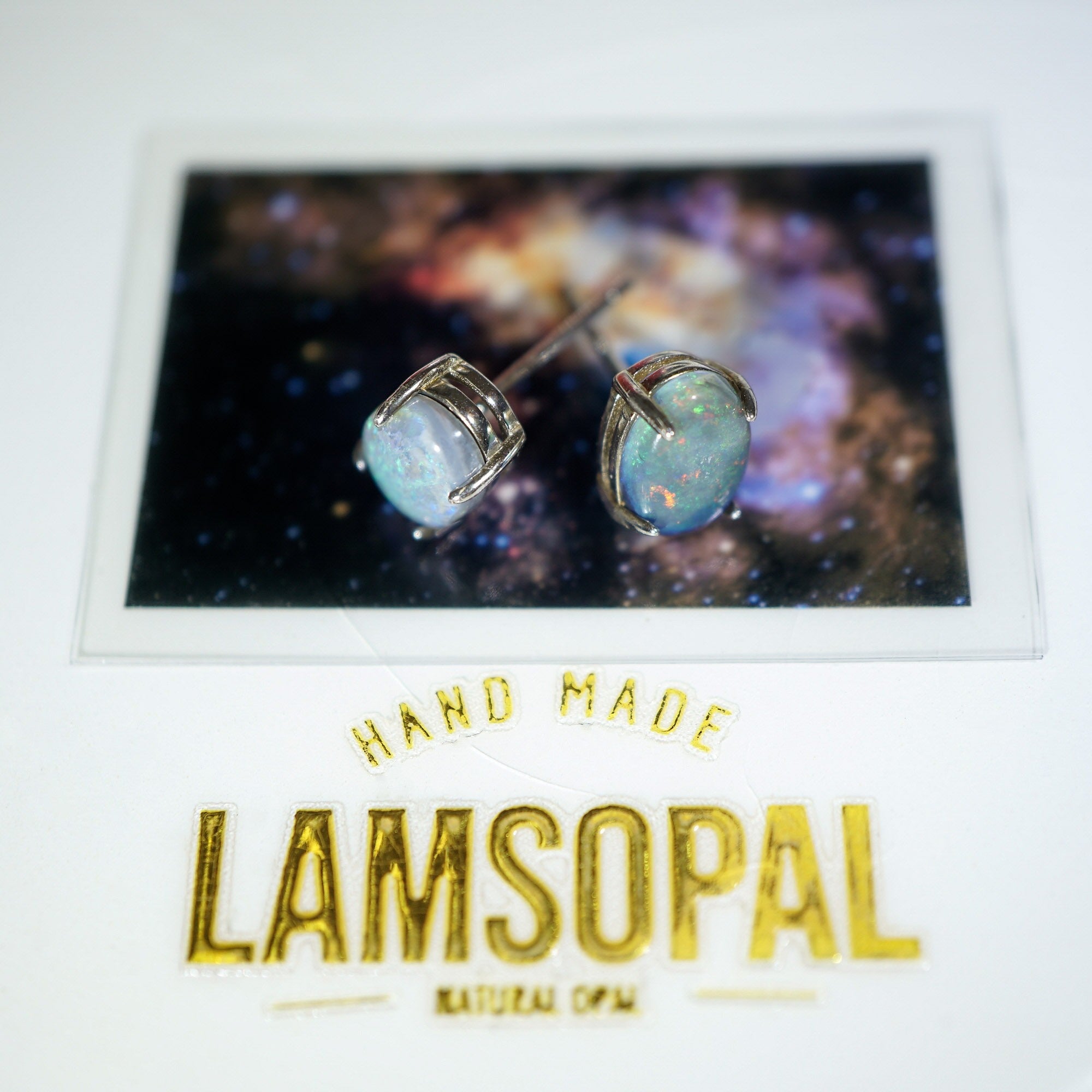 A pair of Lamsopal natural opal stud earrings displayed on a white card. The card features a cosmic galaxy print behind the earrings and gold foil text reading 'HAND MADE LAMSOPAL NATURAL OPAL'.