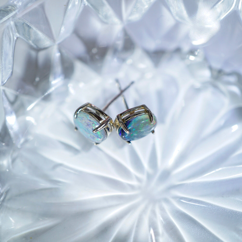 A pair of silver stud earrings with iridescent, opalescent oval-cut stones, resting on a faceted clear glass dish.