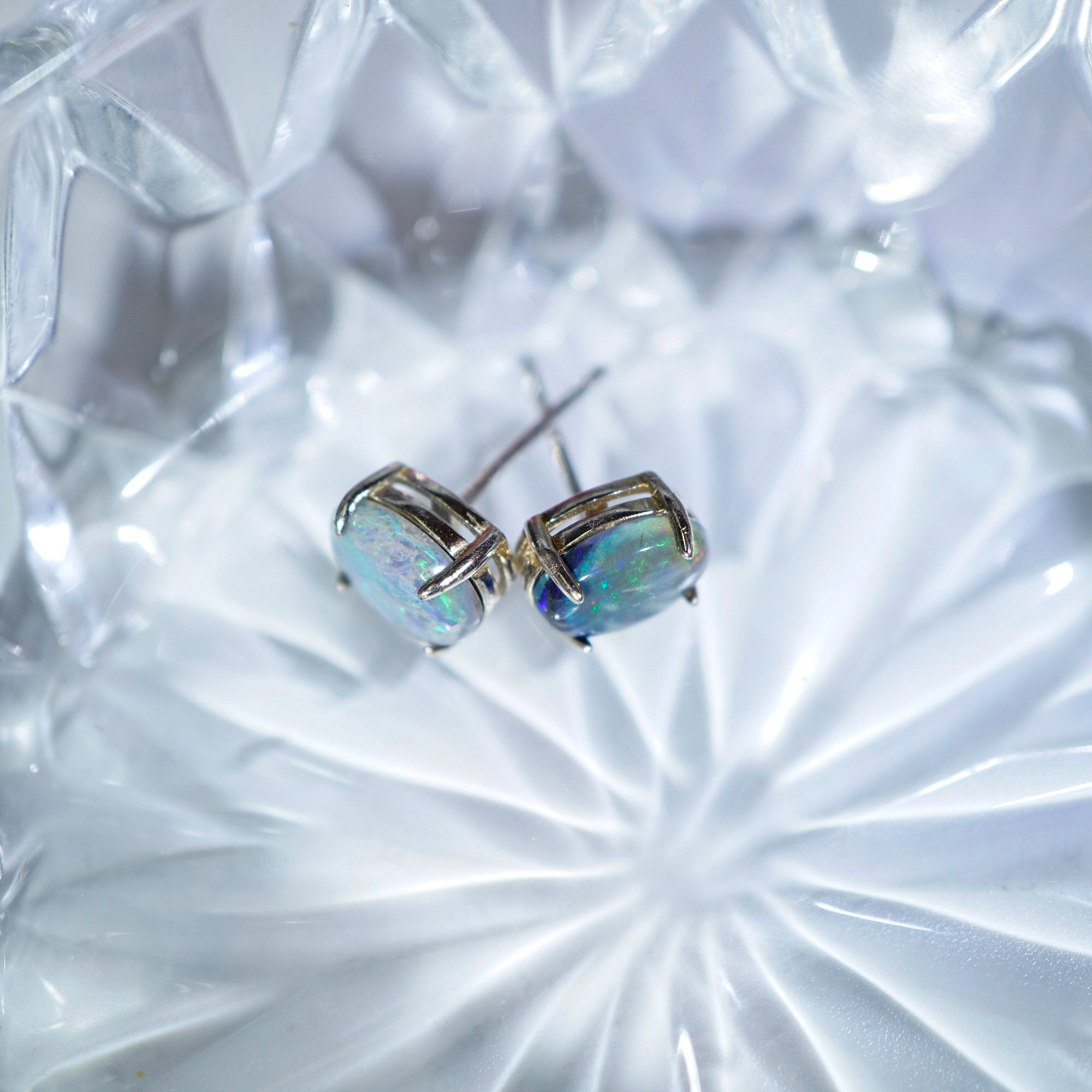 A pair of silver stud earrings with iridescent, opalescent oval-cut stones, resting on a faceted clear glass dish.