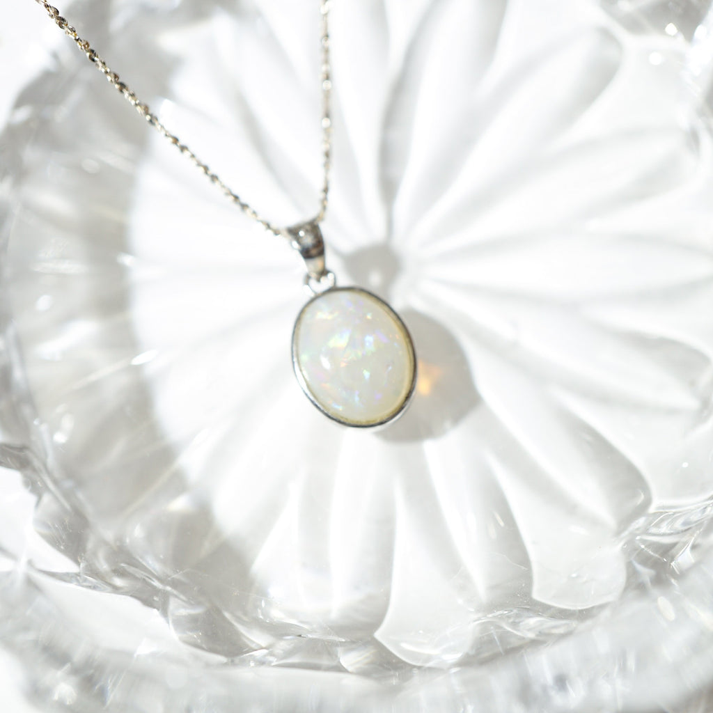 A silver chain necklace with an oval white opal pendant, resting on a clear glass plate.