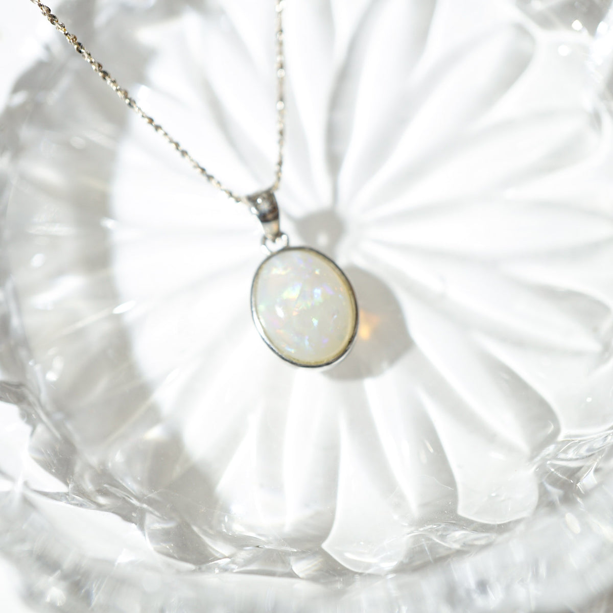A silver chain necklace with an oval white opal pendant, resting on a clear glass plate.